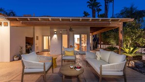 An outdoor patio with two white sofas, a wooden table with a dessert and fruit bowl, and a pergola structure.