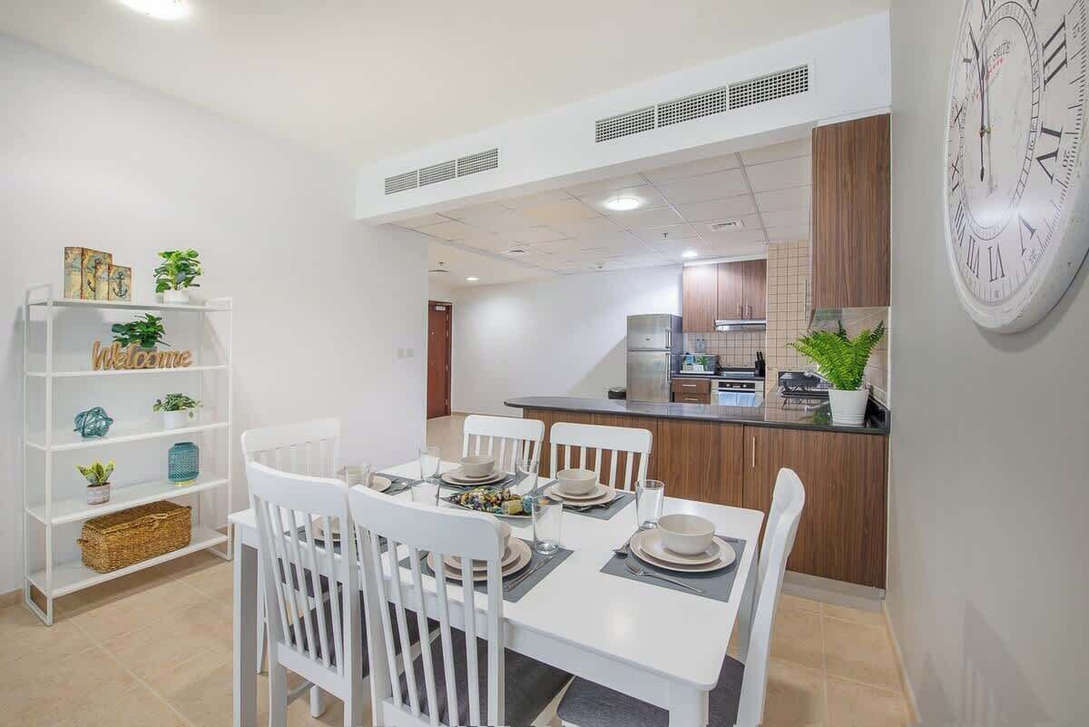 A modern kitchen with white cabinetry and a central island, a dining area with white chairs and a table set for four, and a bookshelf with decorative items.