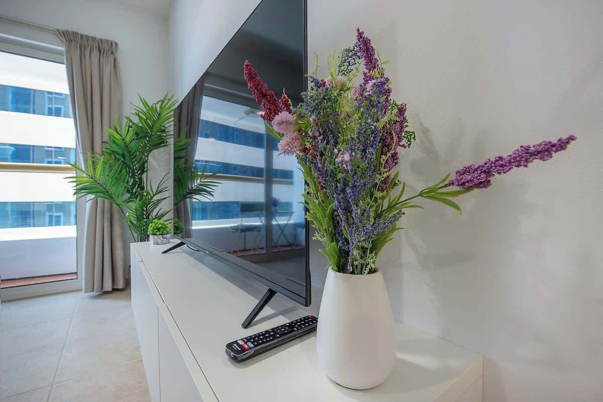 En moderne stue med et fladskærms-tv, en fjernbetjening og en potteplante med lilla blomster.