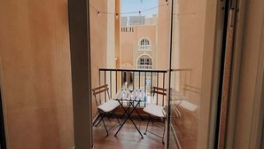 Een smal balkon met een tafel en stoelen, uitzicht op een gebouw met boogvensters, en lichtslingers.