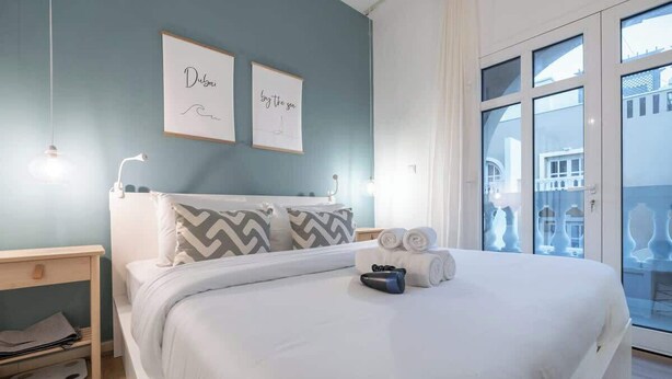 Een opgemaakt bed met witte lakens en een kussen, een nachtkastje met een lamp, en een raam met uitzicht op gebouwen.