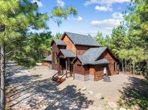 A log cabin with a stone foundation, multiple entry points, and a gravel driveway. - Pop's Place, Pet Friendly, Hot Tub, Play Set, Firewood, and More! (Broken Bow)