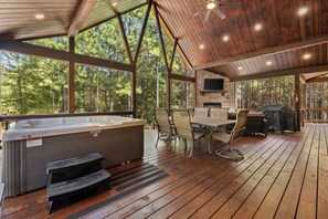A covered outdoor deck with a hot tub, seating area, and a stone fireplace. - Pop's Place, Pet Friendly, Hot Tub, Play Set, Firewood, and More! (Broken Bow)