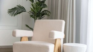 A beige armchair with wooden legs and a matching ottoman in a room with a large plant and sheer curtains. - StayShort - Captivating 1BR apartment in Dubai Creek Harbour! (Dubai)