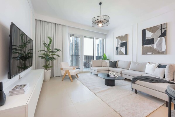 A modern living room with a sofa, coffee table, and a TV. There are plants, a balcony, and artwork on the walls.