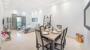 Un comedor moderno con una mesa dispuesta para cuatro personas, rodeada de sillas. Se aprecia un sofá y una mesa de centro al fondo, y una estantería con objetos decorativos.
