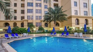 A swimming pool with blue chairs and umbrellas, surrounded by greenery and a multi-story building with arched windows. - StayShort - Modern 2BR Retreat with Breathtaking Views in JBR (Dubai)