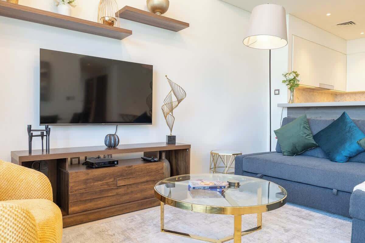 Un salon moderne doté d’un téléviseur à écran plat, d’un meuble de rangement en bois, d’une table basse en verre et d’un canapé gris agrémenté de coussins bleus.