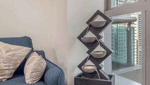 Un salon moderne avec un canapé bleu et des coussins à motifs, un élément décoratif géométrique, et une vue sur des immeubles de grande hauteur par une grande fenêtre.