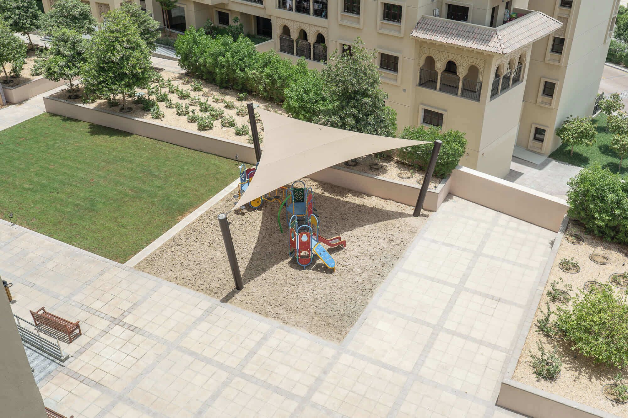 A playground with a slide and climbing structures, surrounded by a landscaped area with trees and a building with arched windows in the background.