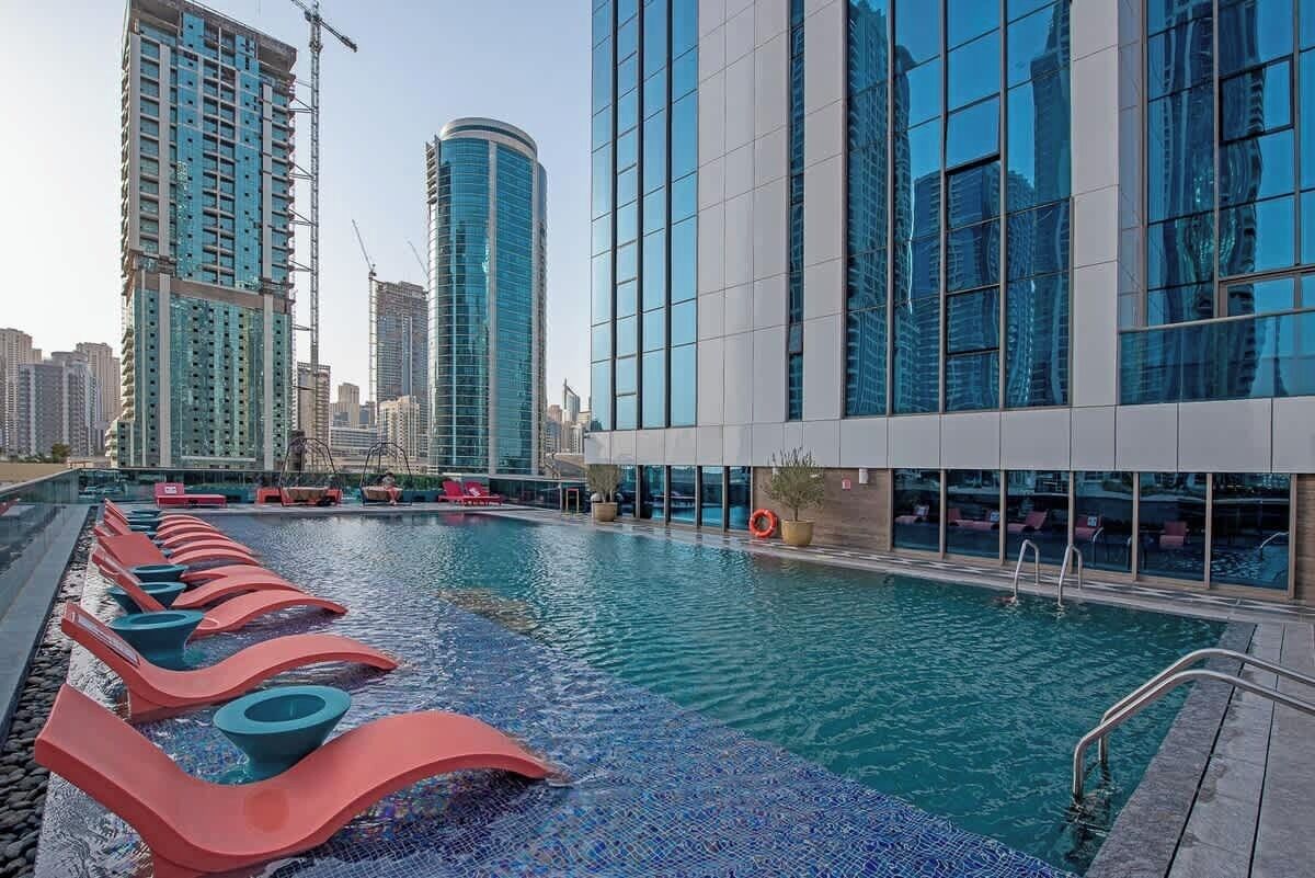 Ein Schwimmbad mit Liegestühlen, ein modernes Gebäude mit großen Fenstern und Hochhäuser im Hintergrund.