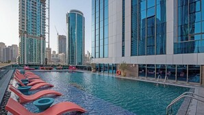 Ein Schwimmbad mit Liegestühlen, ein modernes Gebäude mit großen Fenstern und Hochhäuser im Hintergrund.