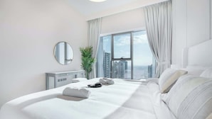 Ein modernes Hotelzimmer mit einem großen Bett, einem runden Spiegel, Blick auf die Stadt durch ein großes Fenster und einer Pflanze.