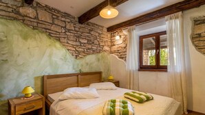 A bedroom with a stone wall, wooden beams, a bed with pillows, a nightstand with a lamp, and a window with curtains. - Villa Prisedi in Rakalj (Rakalj)