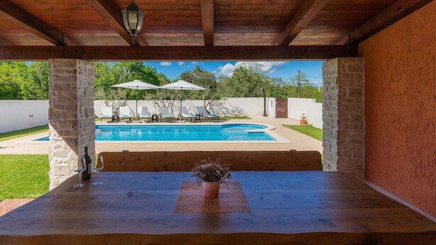 A wooden table with a vase of flowers, a bottle, and glasses, overlooking a swimming pool with lounge chairs and umbrellas. - Villa Prisedi in Rakalj (Rakalj)