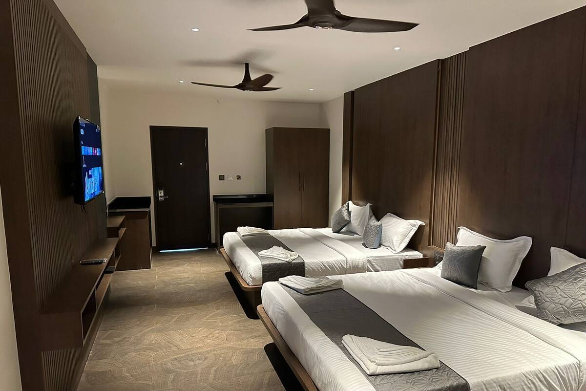 A hotel room with two beds, a TV on the wall, a ceiling fan, and a wooden wardrobe.