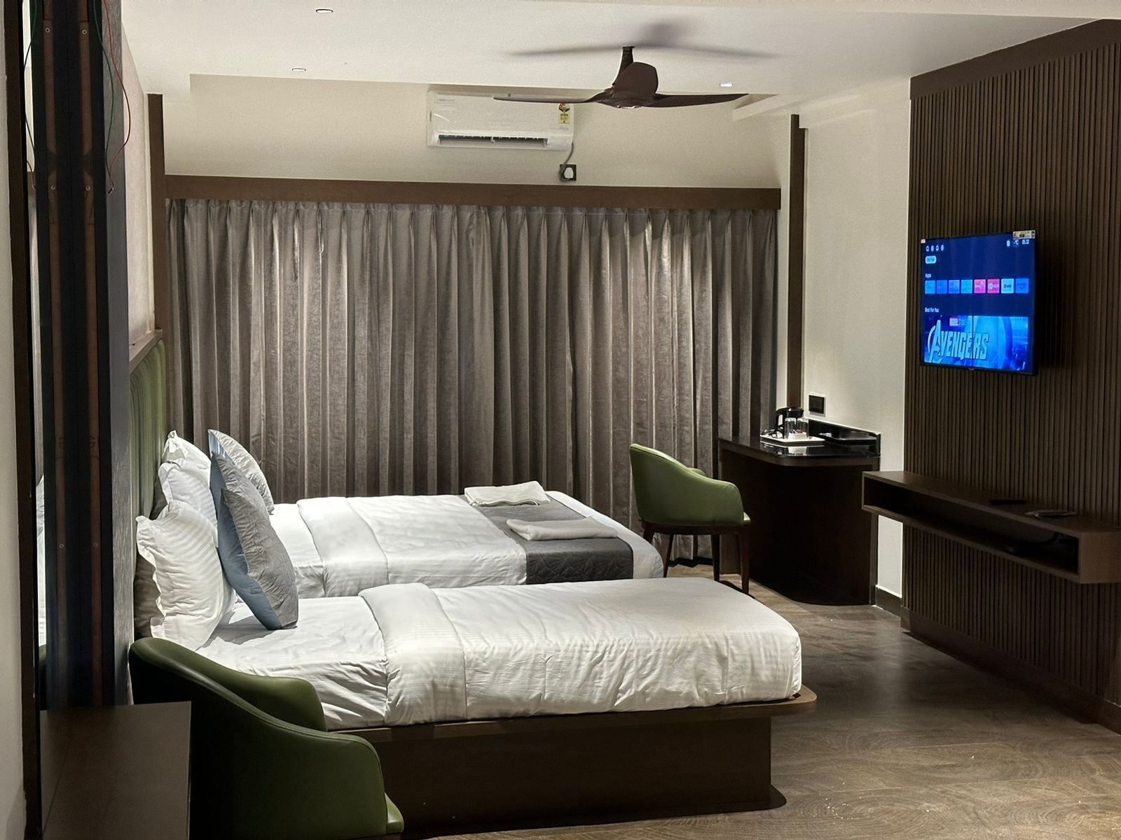 A modern hotel room with a bed, two green armchairs, a desk with a computer, and a TV displaying 'Avengers'.