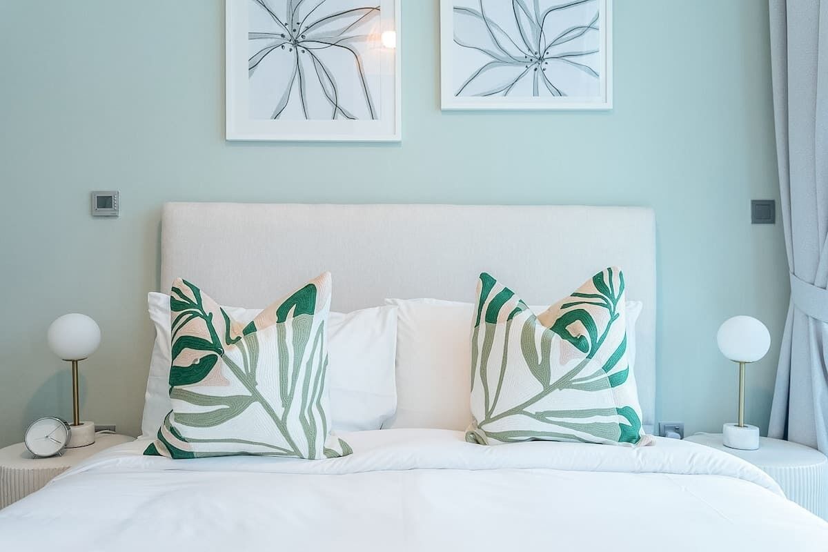 Una cama bien hecha con ropa de cama blanca y dos almohadas decorativas con un estampado de hojas. Encima de la cama hay dos obras de arte colgadas en la pared, y una mesita de noche con una lámpara y un reloj despertador.