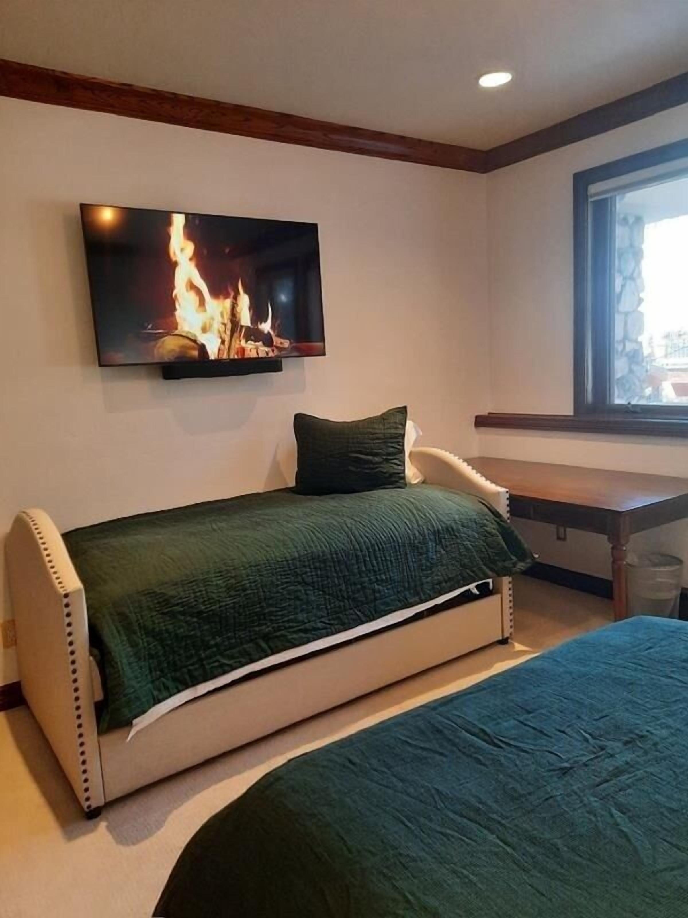 A room with a bed, a TV mounted on the wall displaying a fireplace, a window, and a wooden bench.