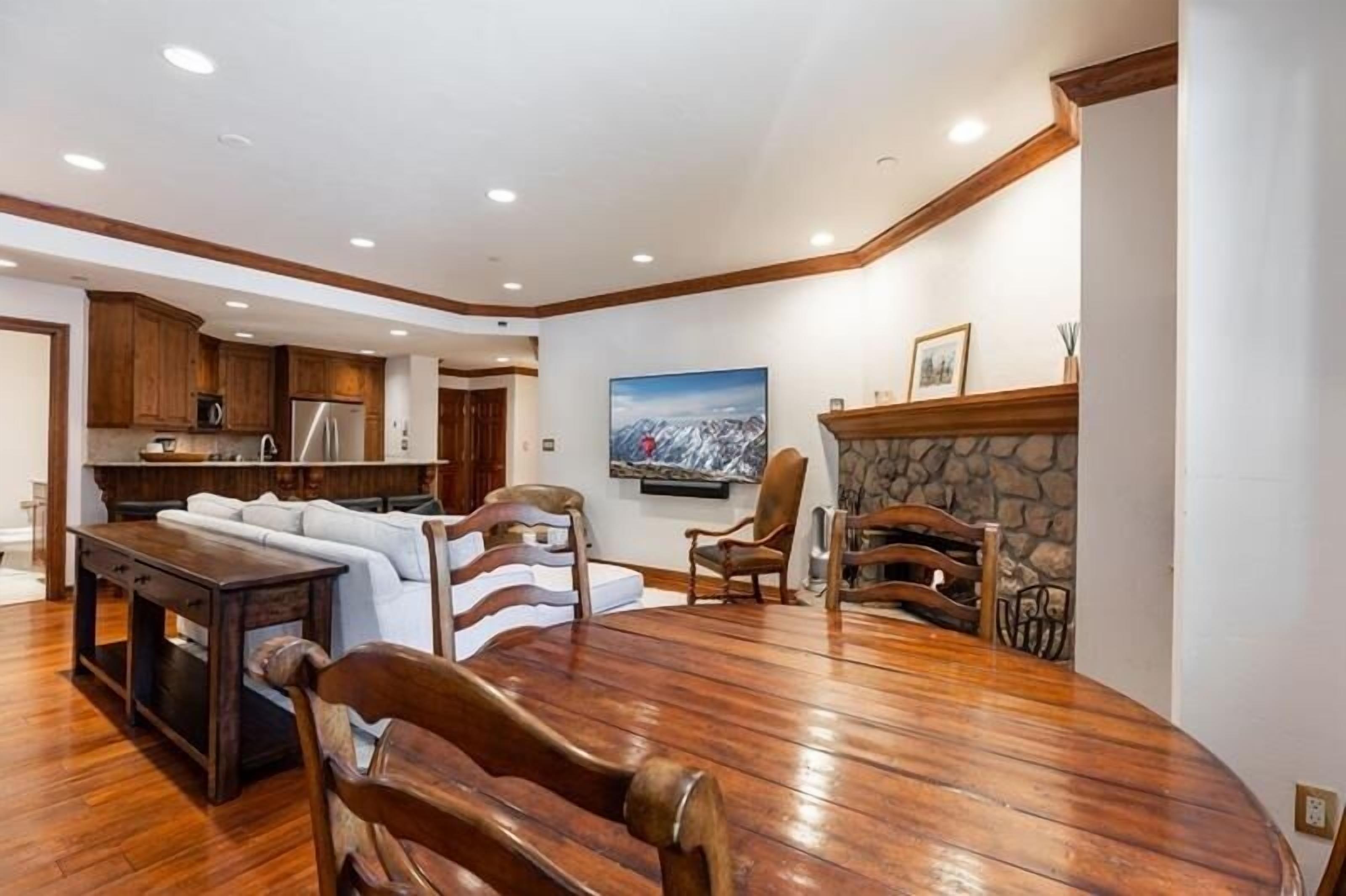 Uma sala de estar ampla com uma mesa de jantar de madeira, lareira e cozinha ao fundo.