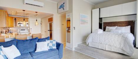 A modern hotel room with a large bed, a blue sofa, a kitchen area, and a bathroom.