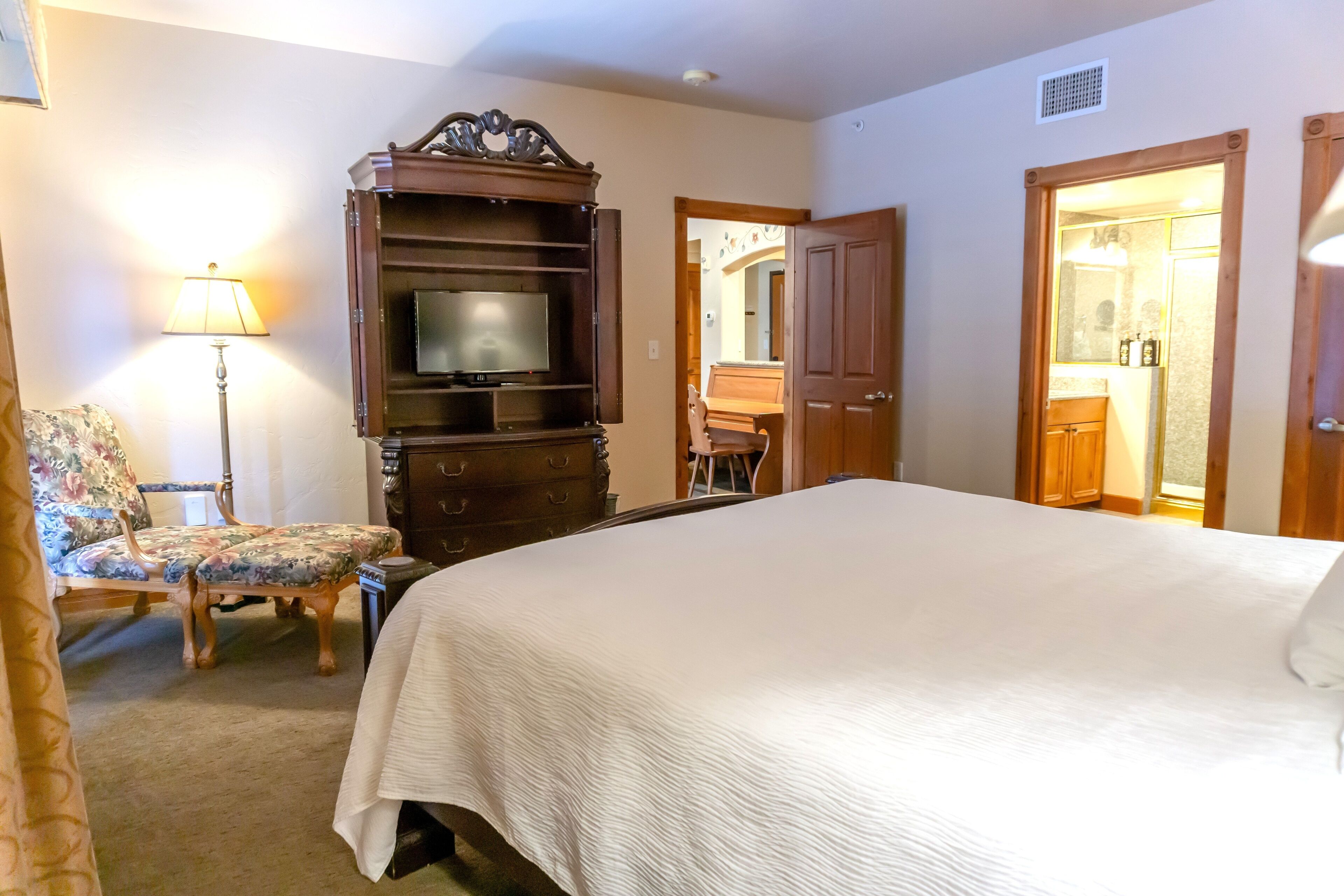 A hotel room with a large bed, a wooden entertainment center with a TV, a floral chair, a desk, and a bathroom visible through an open door.