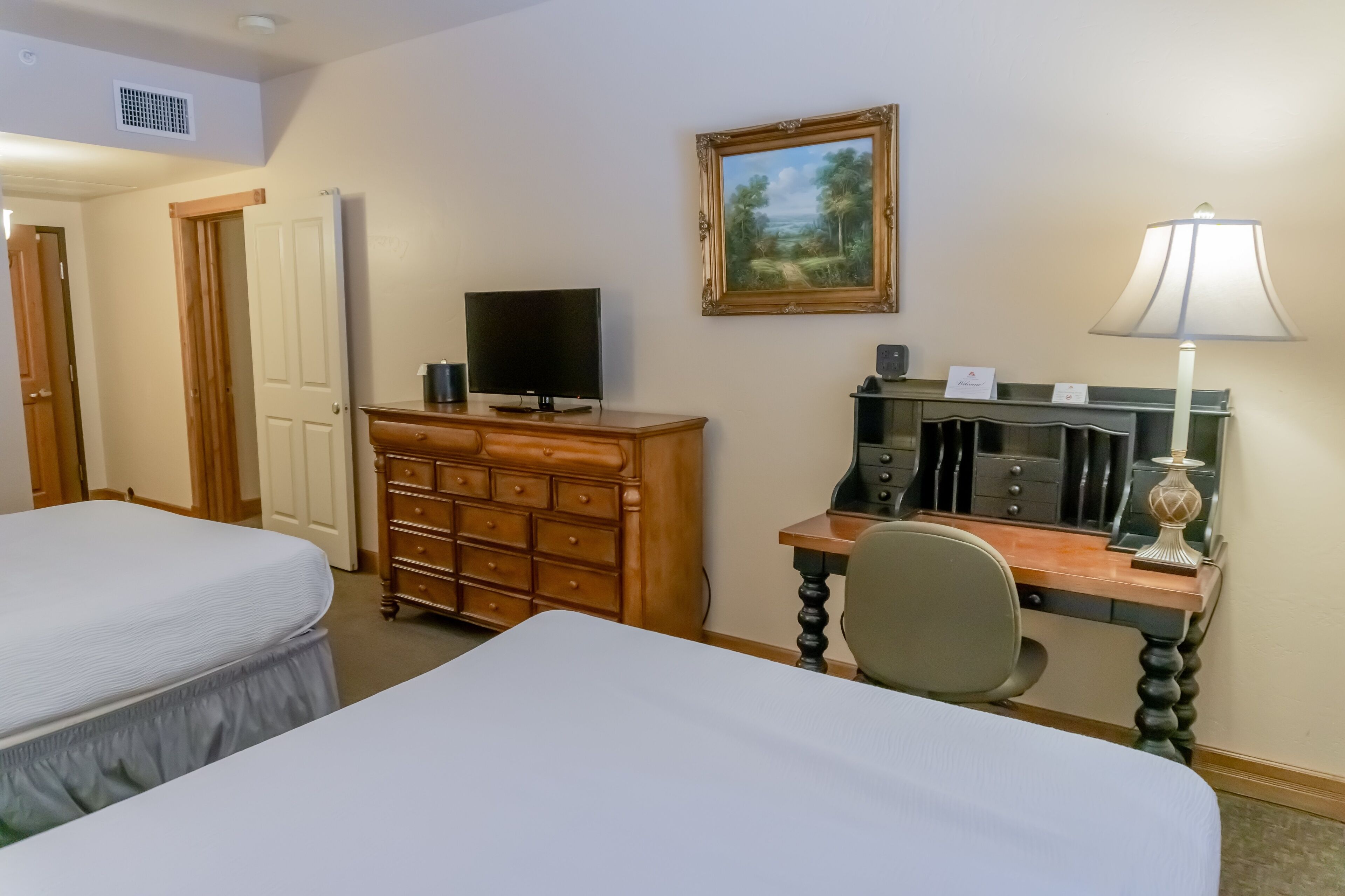 A hotel room with two beds, a wooden dresser, a desk with a lamp, a television, and a framed painting on the wall.