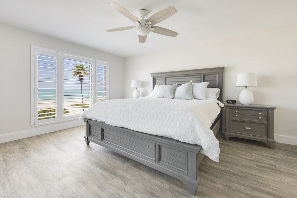 A bedroom with a bed, nightstands, a ceiling fan, and a view of the ocean through large windows.