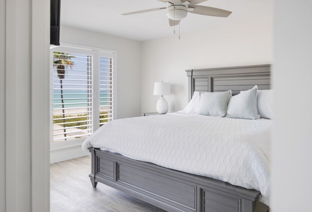 Un dormitorio con una cama grande, un ventilador de techo, una ventana con persianas y vista a la playa.