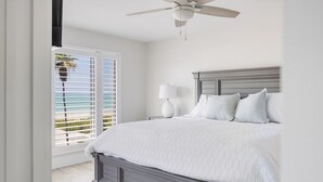 Une chambre avec un grand lit, un ventilateur de plafond, une fenêtre avec des stores et une vue sur la plage.