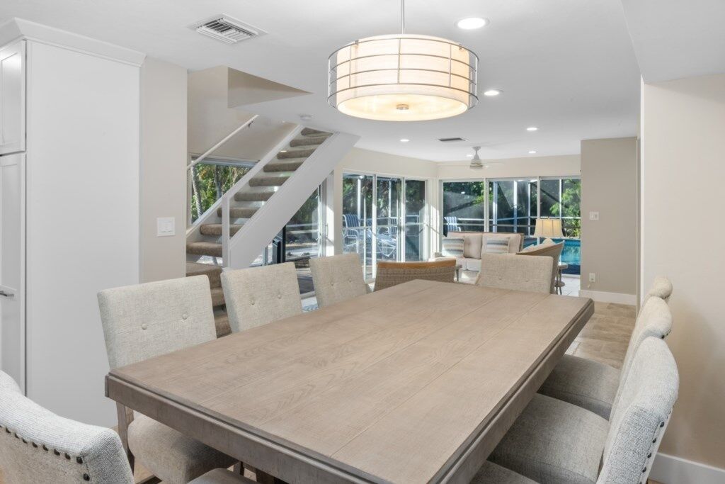 A spacious dining area with a large wooden table and upholstered chairs, a staircase, and large windows offering a view of the outdoors.