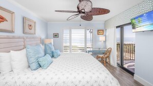 Ein Schlafzimmer mit Bett, Deckenventilator, Schreibtisch und Blick auf den Strand durch große Fenster.