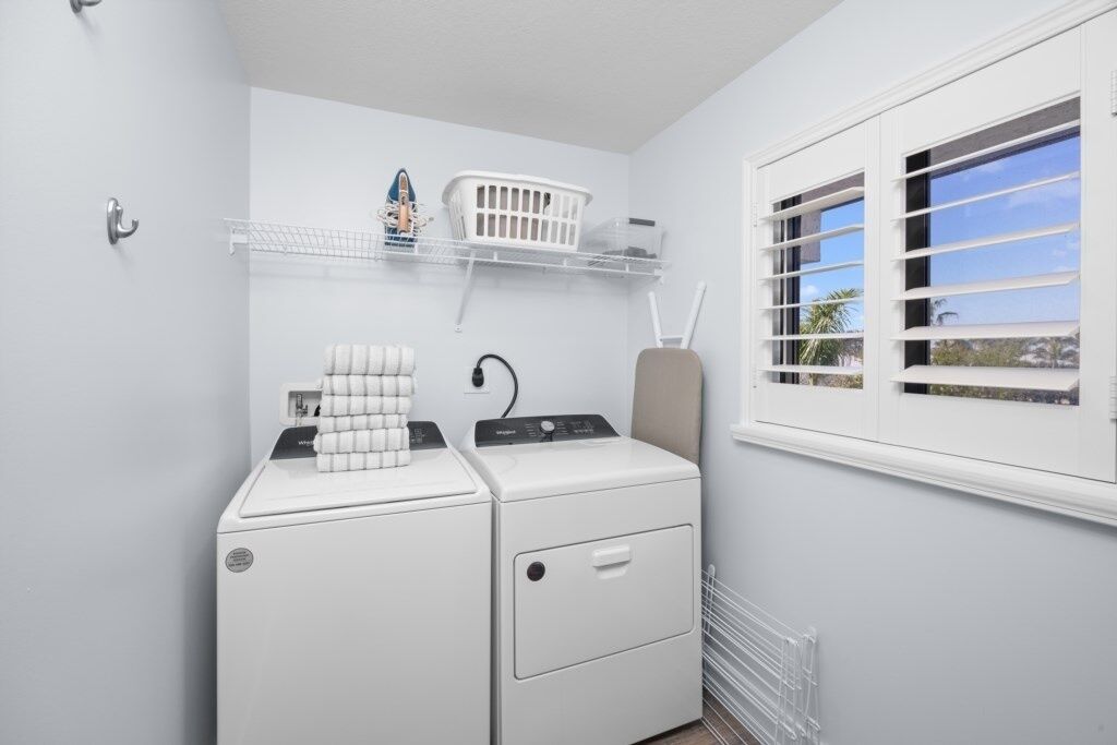 A laundry area with two white washing machines, a drying rack, a shelf with laundry baskets, and a window with blinds.