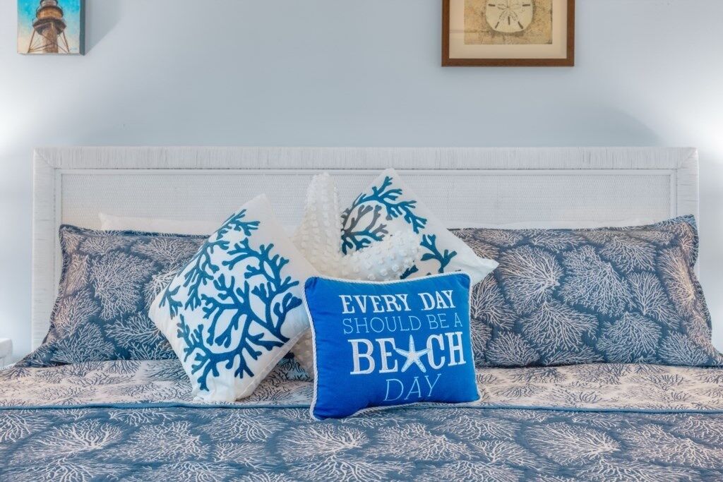 A bed with a white headboard and blue patterned bedding. There are two pillows with blue and white designs, and a blue pillow with the text "EVERY DAY SHOULD BE A BEACH DAY".