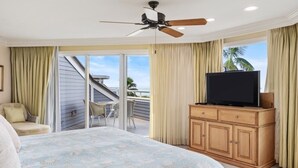 Un dormitorio con cama, televisión sobre un mueble de madera, ventilador de techo y vista al exterior.
