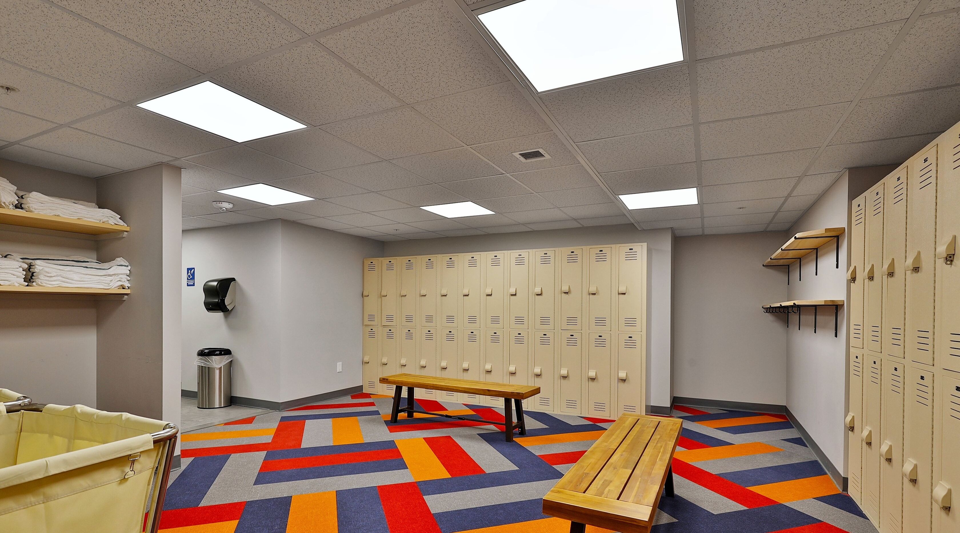 A locker room with multiple lockers, a bench, a trash can, and a colorful floor.