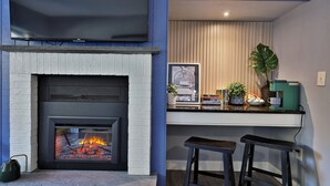 Una sala de estar moderna con chimenea, un televisor de pantalla plana montado arriba y una zona de cocina con taburetes de bar y una encimera.