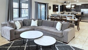 Un salon moderne avec un canapé d’angle, deux petites tables rondes et une cuisine en arrière-plan.