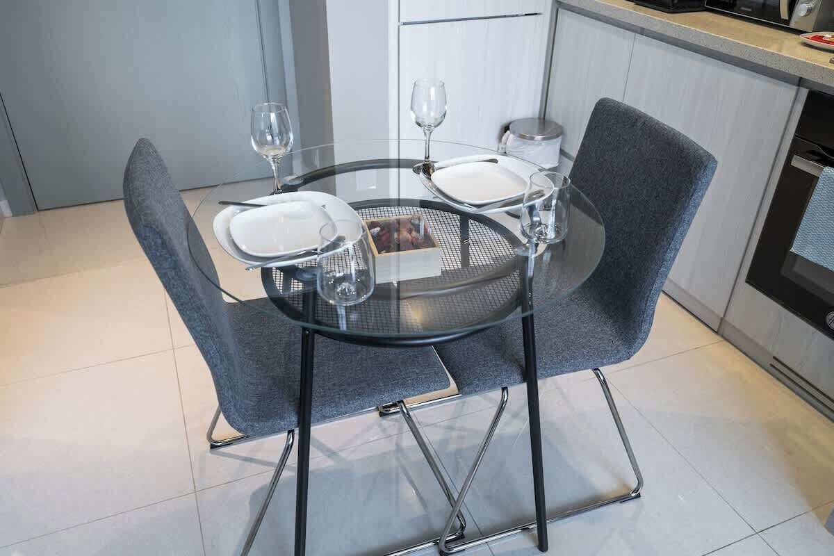 A modern kitchen with a round glass table, grey upholstered chairs, and a white cabinet in the background.