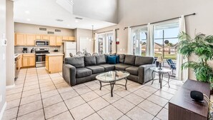A spacious living area with a grey sectional sofa, a glass coffee table, and a dining table with chairs. A kitchen with wooden cabinets and a microwave is visible in the background. - Emerald Island Large 8 Bed Home Golf Resort 8 Bedroom Villa (Kissimmee)
