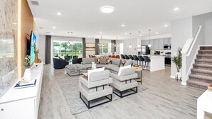 A modern living room with a sectional sofa, a coffee table, and a TV. There is a kitchen area with a dining table and chairs. - Dynamic 15 Bed With Character Bedrooms Pool, Spa! 15 Villa (Kissimmee)