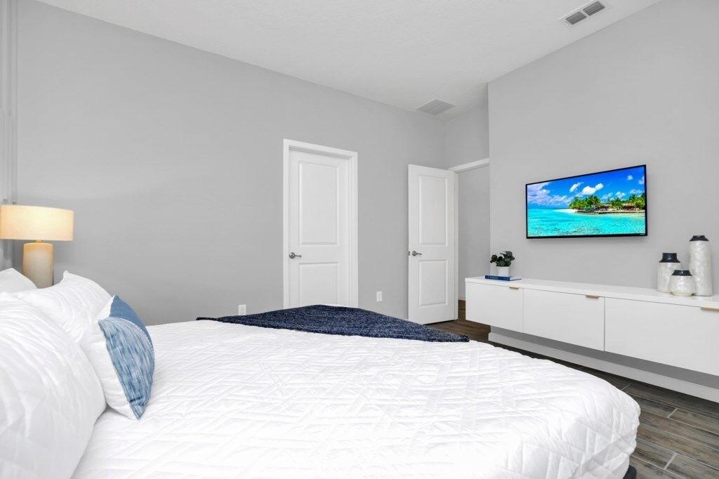 Ein Schlafzimmer mit einem Bett, einem an der Wand montierten Fernseher, einem Nachttisch mit Lampe und einer Tür.