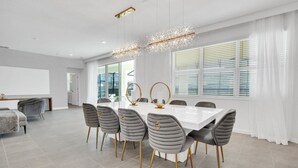 Une salle à manger moderne avec une table blanche et des chaises grises à revêtement, de grandes fenêtres équipées de stores et un lustre.