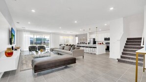 Un salon moderne avec un canapé, une table basse et un coin repas avec des chaises. Au fond, on aperçoit une cuisine et un escalier menant à un autre niveau.