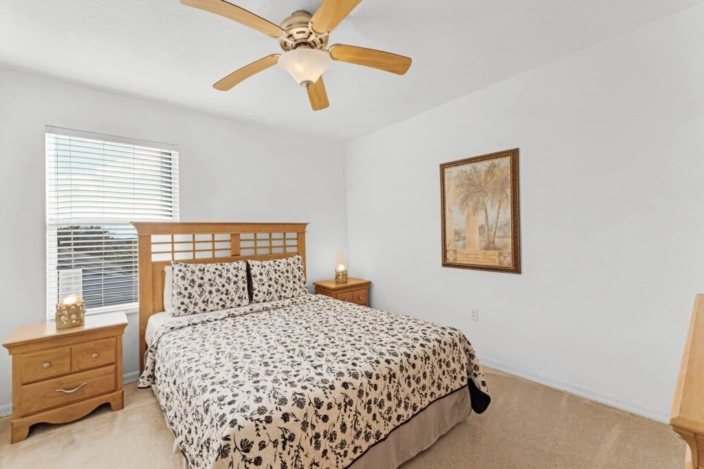 A bedroom with a wooden bed, a nightstand with a lamp, a ceiling fan, a window with blinds, and a framed picture on the wall.