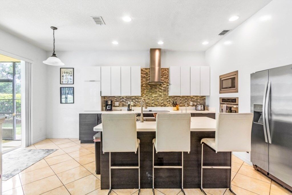 A modern kitchen with a central island, white cabinets, a stainless steel refrigerator, and a stone backsplash.