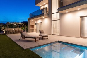 Un área de piscina moderna al aire libre con dos sillones de descanso y una pérgola con luces colgantes.