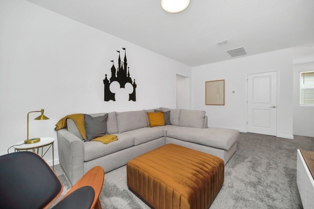 En moderne stue med en grå hjørnesofa, en brun skammel og en sort lænestol. Der er en sort vægdekal af et slot, et lille rundt bord med en lampe og et indrammet billede på væggen.