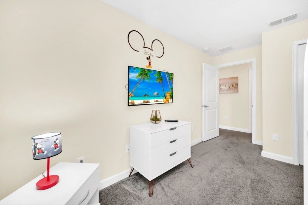 Une chambre avec une commode blanche, un autocollant mural à l’effigie de Mickey Mouse, un téléviseur diffusant une scène de plage et une lampe décorée avec le motif de Mickey Mouse.