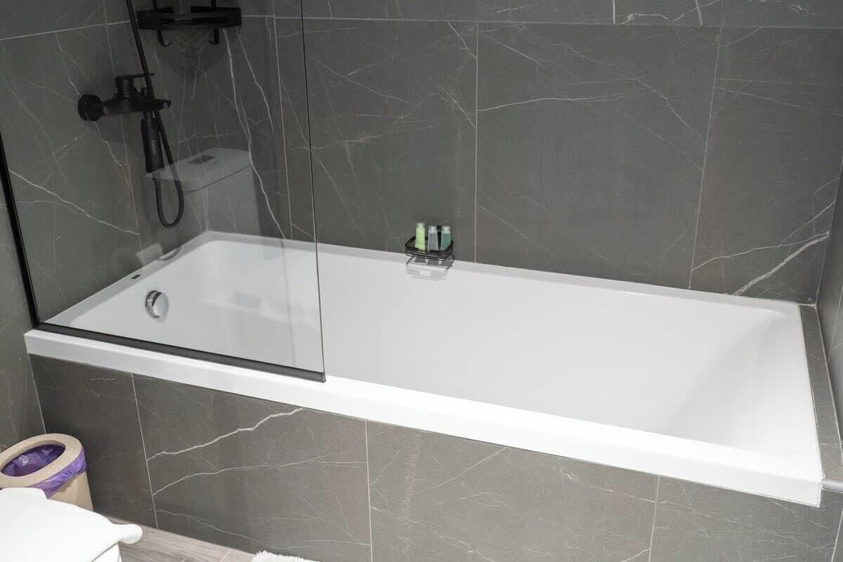Et moderne badeværelse med et hjørnebadekar med glasvægge, et håndholdt brusehoved og en lille hylde med toiletartikler.